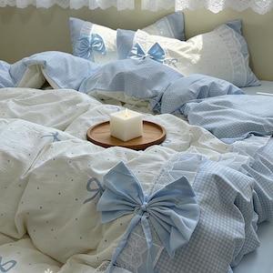 Puede incluir: Un juego de cama azul claro y blanco con diseño de lazo. El juego incluye una funda nórdica, almohadas y una sábana. La funda nórdica tiene un estampado de cuadros y un adorno de lazo. Las almohadas tienen un lazo y ribete de encaje. Una bandeja de madera con una vela blanca está sobre la cama.