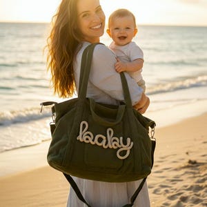 Może przedstawiać: Oliwkowo-zielona torba dla dziecka z napisem „baby” w białym skrypcie. Torba ma pasek na ramię i jest trzymana przez osobę na plaży. Dziecko jest trzymane przez tę osobę.