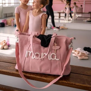 Peut inclure: Un sac fourre-tout en velours côtelé rose avec le mot "mama" en écriture blanche. Le sac a une bandoulière et un charm de ballerine. Deux jeunes filles en justaucorps roses se tiennent devant le sac dans un studio de danse.