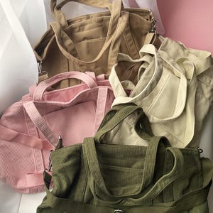 May include: A collection of corduroy tote bags in various colors: tan, pink, cream, and olive green. Each bag features a structured design with top handles and a shoulder strap. The bags are arranged in a pile, showcasing their different hues and textures.