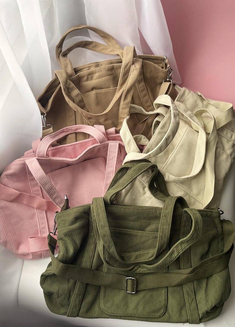 May include: A collection of corduroy tote bags in various colors: tan, pink, cream, and olive green. Each bag features a structured design with top handles and a shoulder strap. The bags are arranged in a pile, showcasing their different hues and textures.