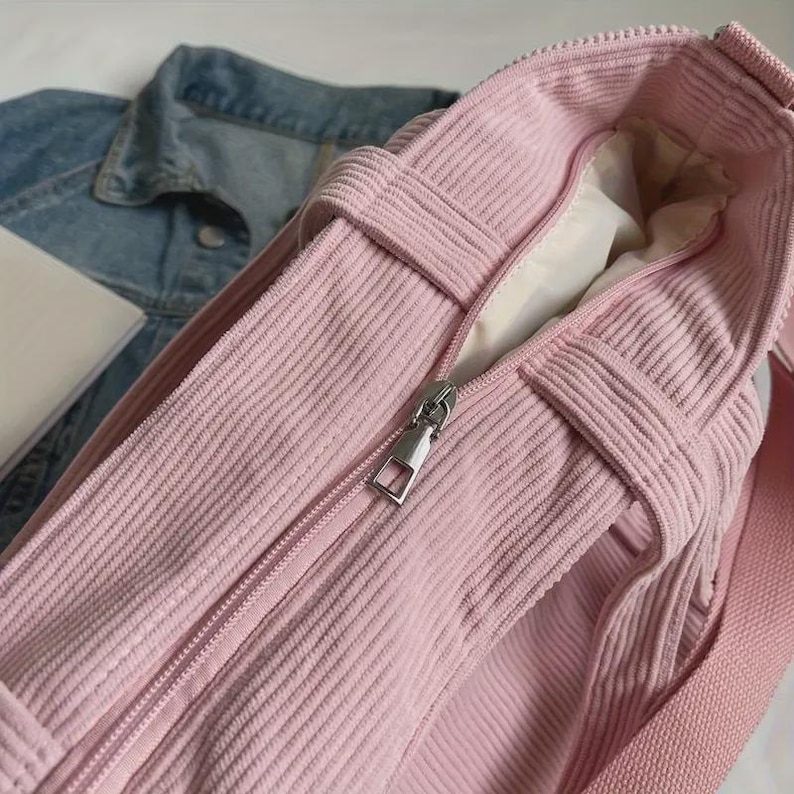 May include: A close-up of a pink corduroy tote bag with a silver zipper. The bag has a rectangular shape with a long shoulder strap. A denim jacket is visible in the background.