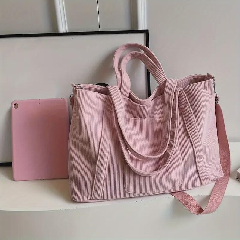 May include: A light pink corduroy tote bag with a matching shoulder strap and handles. The bag has a rectangular shape with a front pocket and a zipper closure. A pink tablet is next to the bag.