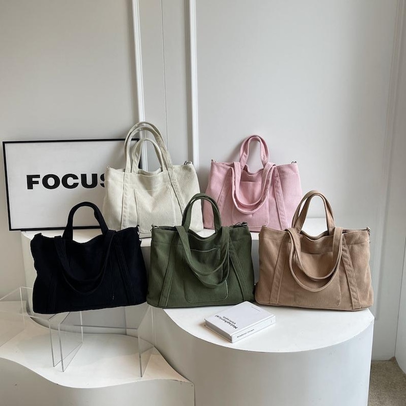 May include: A collection of corduroy tote bags in various colors: black, cream, pink, olive green, and tan. Each bag has a rectangular shape with two handles and a shoulder strap. The word "FOCUS" is visible on a sign in the background.