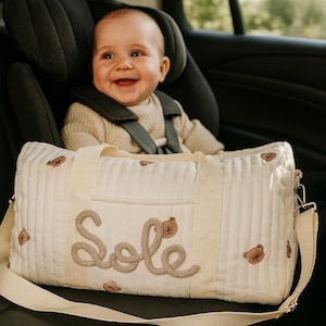 May include: Cream-colored baby bag with a quilted design and embroidered teddy bears. The word "Sole" is stitched in a cursive font on the front. The bag has a shoulder strap and a top handle. A baby is in a car seat in the background.