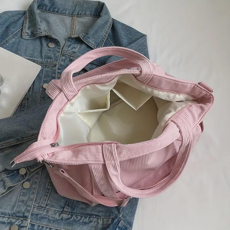 May include: A pink corduroy tote bag with a zipper closure and two handles. The interior features multiple pockets and a white lining. The bag is shown open, revealing the inside. The bag is a light pink color.
