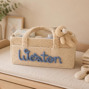 May include: A beige baby diaper caddy with the name "Weston" in blue script. The caddy has a teddy bear on top and contains diapers and other baby essentials. A pair of beige baby booties and a stack of folded cloths are nearby.