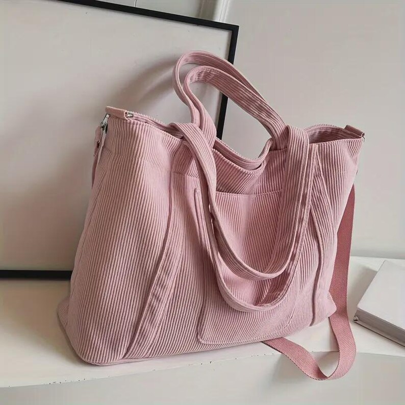 May include: A pink corduroy tote bag with a shoulder strap and two handles. The bag has a zippered top and an exterior pocket. The bag is sitting on a white surface.