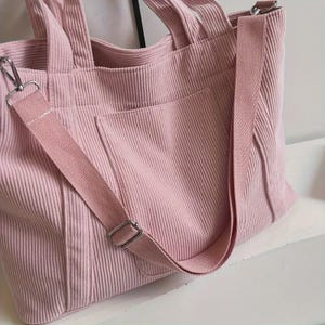 May include: A pink corduroy tote bag with a matching adjustable shoulder strap. The bag has a front pocket and silver-tone metal hardware. The tote bag is designed with a structured shape and a soft, textured corduroy fabric.