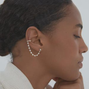May include: A gold ear cuff featuring a chain of small, clear gemstones. The ear cuff wraps around the upper ear and connects to the chain, which hangs down. The jewellery is set against a neutral background, highlighting the design.