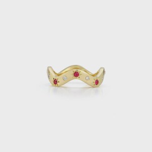 Puede incluir: Un anillo dorado con un diseño ondulado. El anillo está adornado con pequeñas piedras preciosas rojas en forma de estrella y pequeñas piedras preciosas redondas y claras. El diseño del anillo es simétrico, con las piedras espaciadas.