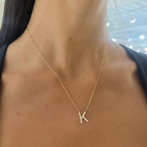 Pode incluir: Um colar dourado com uma corrente delicada. O pingente é a letra "K" incrustada com pequenas pedras brilhantes. O colar é usado em volta do pescoço, com o pingente repousando no peito.