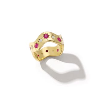 Könnte beinhalten: Ein goldfarbener Ring mit wellenförmigem Design. Der Ring ist mit abwechselnd roten Edelsteinen und kleinen weißen Edelsteinen verziert. Der Ring hat ein Sternmuster um die roten Edelsteine. Auf weißem Hintergrund.