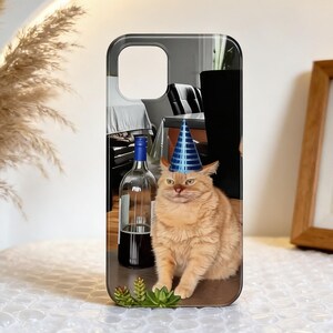 May include: A phone case with a ginger cat wearing a blue party hat. The cat is in front of a wine bottle. The case has a glossy finish and a camera cutout.