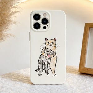 May include: A white phone case featuring a whimsical illustration of two cats embracing. The top cat is light orange, and the bottom cat is gray and white. The case has a triple-lens camera cutout and a flash.