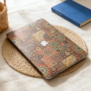 May include: A laptop with a colorful, patterned cover featuring a variety of floral and geometric designs in shades of red, orange, green, and tan. The laptop sits on a round, woven placemat. A blue book is in the background.