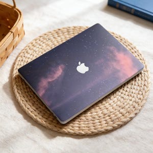 May include: A closed laptop with a starry sky design on a woven mat. The laptop has a white Apple logo. The background features a woven basket and a blue book.