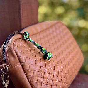 Op de afbeelding: Een bruine geweven leren etui met een zwarte ritssluiting en een zilverkleurige sluiting. Een groen, blauw en zwart touw is aan de ritssluiting bevestigd. De etui is rechthoekig en heeft een gestructureerd oppervlak.