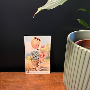 Peut inclure: Une carte postale vintage avec une illustration d'un garçon et d'une fille, accompagnés d'un petit chien. Le garçon porte une chemise rayée et un chapeau, la fille a un nœud rose. Le texte en bas dit "THANK HEAVEN - FOR LITTLE GIRLS!"