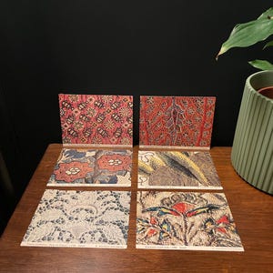 May include: Six vintage postcards with colorful, intricate patterns are displayed on a wooden surface. The postcards feature floral and abstract designs in shades of red, blue, yellow, and white. A green potted plant is in the background.