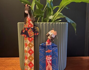 May include: Two origami geisha dolls stand in front of a green plant in a ribbed pot. The dolls are made of patterned paper in red, blue, and gold. One doll is taller and has a red and gold patterned kimono. The other doll is shorter and has a blue kimono.