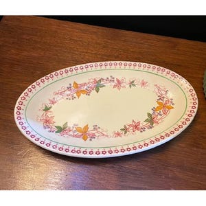 Könnte beinhalten: Ovale Servierplatte mit cremefarbenem Mittelteil und dekorativem Rand. Der Rand weist ein sich wiederholendes Muster aus kleinen roten Blüten und grünen Blättern auf. Die Mitte hat ein Blumendesign mit orangefarbenen und rosa Blättern.