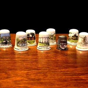 May include: A selection of decorative thimbles, each showcasing a different design. The thimbles depict various landmarks and scenes, including buildings and landscapes. One thimble is metallic with an anchor design. The thimbles are displayed on a wooden surface.