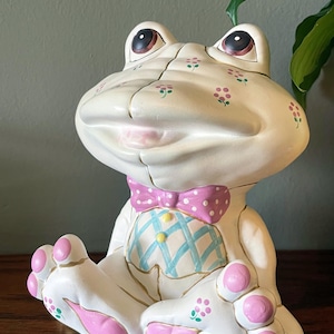 May include: A ceramic frog figurine in a seated position. The frog is primarily off-white with pink accents on the feet and a pink polka-dot bow tie. The frog's chest has a blue and white plaid pattern. Small pink flower details are scattered across the body.
