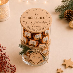 Könnte beinhalten: Ein klares Glasgefäß gefüllt mit einzeln verpackten Pralinen, verschlossen mit einem runden Holzdeckel, der mit 24 KÜSSCHEN bis Weihnachten für Vanessa graviert ist, sowie einem kleinen Holzanhänger mit der Aufschrift Für dich an einer Kordel.