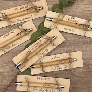 Könnte beinhalten: Eine Auswahl an personalisierten Bambusstiften auf Holzschildern, mit Kordel gebunden. Jeder Stift hat eine silberne Spitze und ist mit einem Namen und einer kurzen Botschaft graviert. Die Karten tragen Aufschriften wie "Für meinen Lieblingsmenschen".