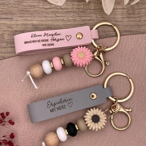 Könnte beinhalten: Zwei Schlüsselanhänger mit goldfarbener Hardware. Einer ist rosa mit dem Text "Kleine Menschen brauchen so große Herzen wie deins" und einem Gänseblümchen-Charm. Der andere ist grau mit dem Text "Erzieherin mit Herz" und einem Sonnenblumen-Charm. Beide haben Perlenakzente.