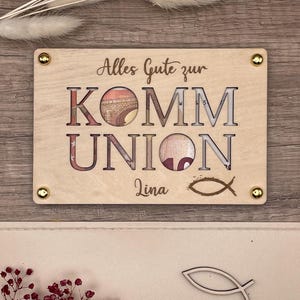 May include: A light-colored wooden plaque with the words "Alles Gute zur KOMM UNION" cut out, revealing currency. The name "Lina" and a fish symbol are below. Gold-colored screws secure the plaque.