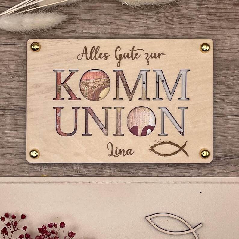 May include: A light-colored wooden plaque with the words "Alles Gute zur KOMM UNION" cut out, revealing currency. The name "Lina" and a fish symbol are below. Gold-colored screws secure the plaque.