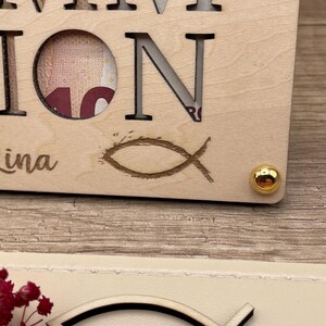 May include: A wooden plaque with the word "ION" and the name "ina" is visible. The letter "O" has a circular cutout revealing a portion of a bill. A fish symbol is etched below the letter "N". A gold-colored stud is in the corner.