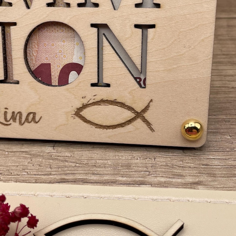 May include: A wooden plaque with the word "ION" and the name "ina" is visible. The letter "O" has a circular cutout revealing a portion of a bill. A fish symbol is etched below the letter "N". A gold-colored stud is in the corner.