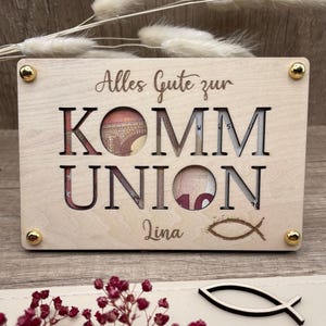 May include: A light-colored wooden gift box with the words "Alles Gute zur KOMMUNION" cut out. The name "Lina" and a fish symbol are engraved below. Gold-colored studs secure the corners.