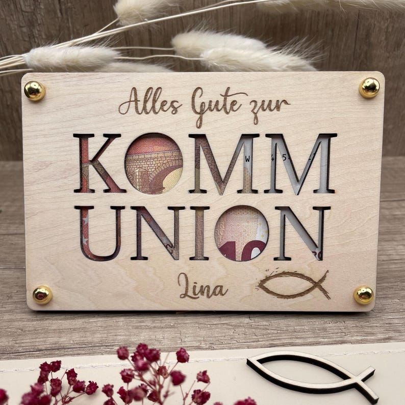 May include: A light-colored wooden gift box with the words "Alles Gute zur KOMMUNION" cut out. The name "Lina" and a fish symbol are engraved below. Gold-colored studs secure the corners.