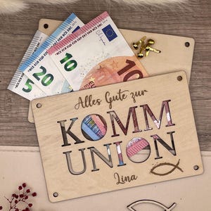 May include: A wooden gift box with the words "Alles Gute zur KOMM UNION" cut out, with euro banknotes visible through the cut-out letters. The box also has a small fish symbol and the name "Lina".