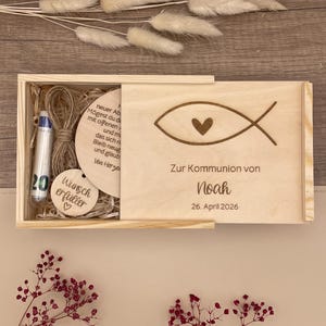 Könnte beinhalten: Eine Holzkiste mit Schiebedeckel, graviert mit einem Fischsymbol und einem Herz. Der Text "Zur Kommunion von Noah 26. April 2026" ist sichtbar. Im Inneren befinden sich ein Stift, eine Holzscheibe und ein Anhänger mit "Wunsch erfüller".