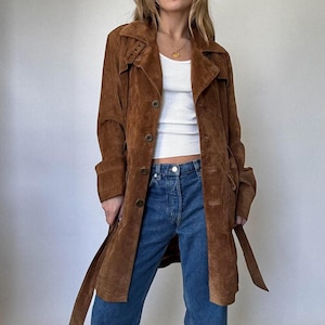 Könnte beinhalten: Ein brauner Wildleder-Trenchcoat mit Knopfleiste, Gürtel und klassischem Kragen. Der Mantel ist knielang und hat lange Ärmel. Das Model trägt eine blaue Jeans und ein weißes Tanktop. Der Mantel hat eine mittelbraune Farbe.