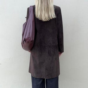 May include: A dark brown suede coat with a matching large shoulder bag in a deep purple color. The coat is knee-length and has a classic design. The bag is draped over the shoulder.