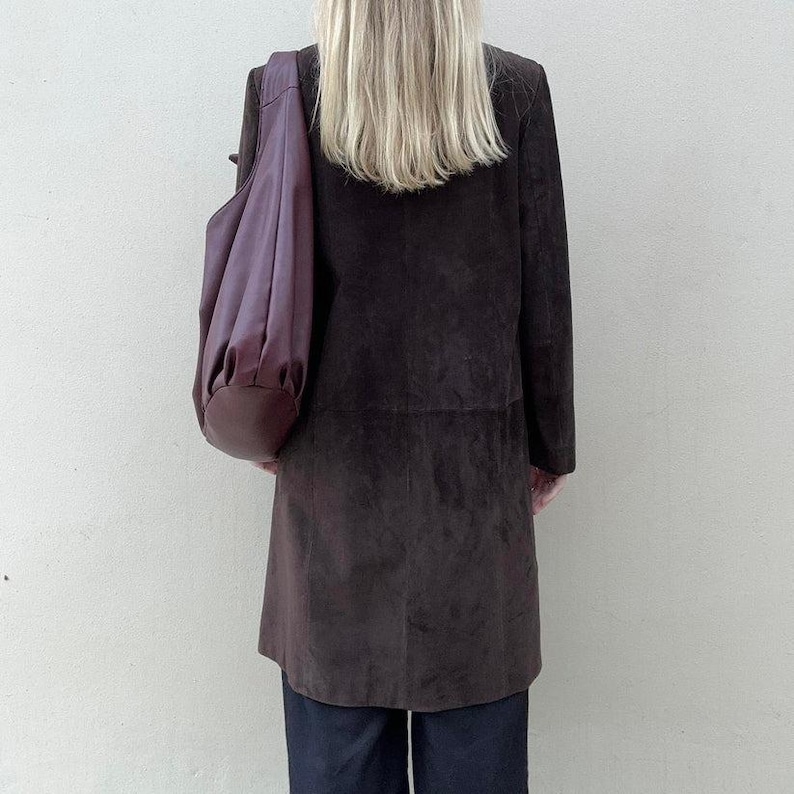 May include: A dark brown suede coat with a matching large shoulder bag in a deep purple color. The coat is knee-length and has a classic design. The bag is draped over the shoulder.