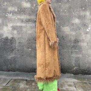 May include: A long, tan suede coat with faux fur trim on the collar, cuffs, and hem. The coat is worn over bright green pants. The coat has a classic, vintage style.