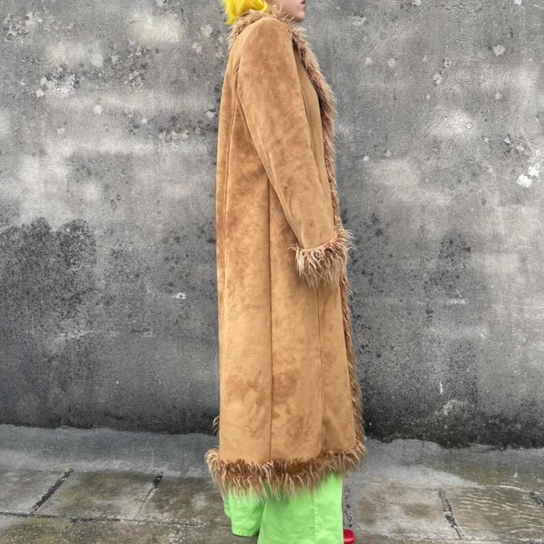 May include: A long, tan suede coat with faux fur trim on the collar, cuffs, and hem. The coat is worn over bright green pants. The coat has a classic, vintage style.