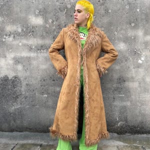 May include: A long, tan suede coat with faux fur trim around the collar, cuffs, and hem. The coat is open in the front, revealing a green shirt and pants. The coat has a vintage, bohemian style.
