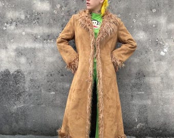 May include: A long, tan suede coat with faux fur trim around the collar, cuffs, and hem. The coat is open in the front, revealing a green shirt and pants. The coat has a vintage, bohemian style.