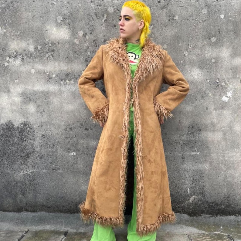 May include: A long, tan suede coat with faux fur trim around the collar, cuffs, and hem. The coat is open in the front, revealing a green shirt and pants. The coat has a vintage, bohemian style.