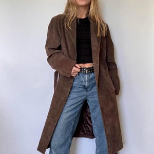 May include: A long, brown suede coat with a button-down front and a matching belt. The coat has a classic lapel collar and falls to the ankles. The coat is worn over a black top and blue jeans.