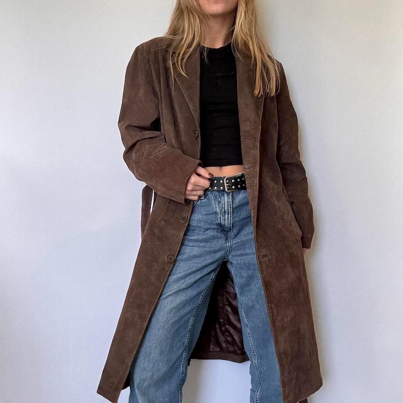 May include: A long, brown suede coat with a button-down front and a matching belt. The coat has a classic lapel collar and falls to the ankles. The coat is worn over a black top and blue jeans.