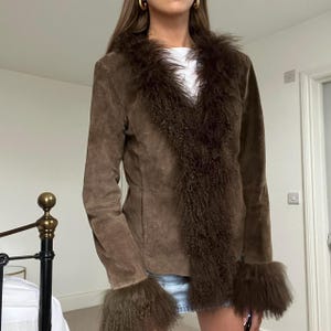 Puede incluir: Chaqueta de ante marr&oacute;n con cuello y pu&ntilde;os de piel sint&eacute;tica. La chaqueta tiene cuello en V y mangas largas. El ribete de piel sint&eacute;tica es de un marr&oacute;n m&aacute;s oscuro, creando contraste con el ante. Una prenda de abrigo elegante.